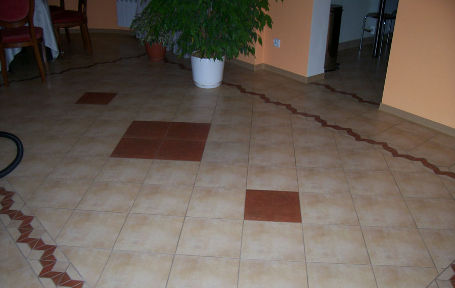 Beżowa podłoga z płytek ceramicznych z wkomponowanymi kwadratami w kolorze brązowym i dekoracyjnym bordiurem w kształcie zygzaka, w tle doniczka z zieloną rośliną.