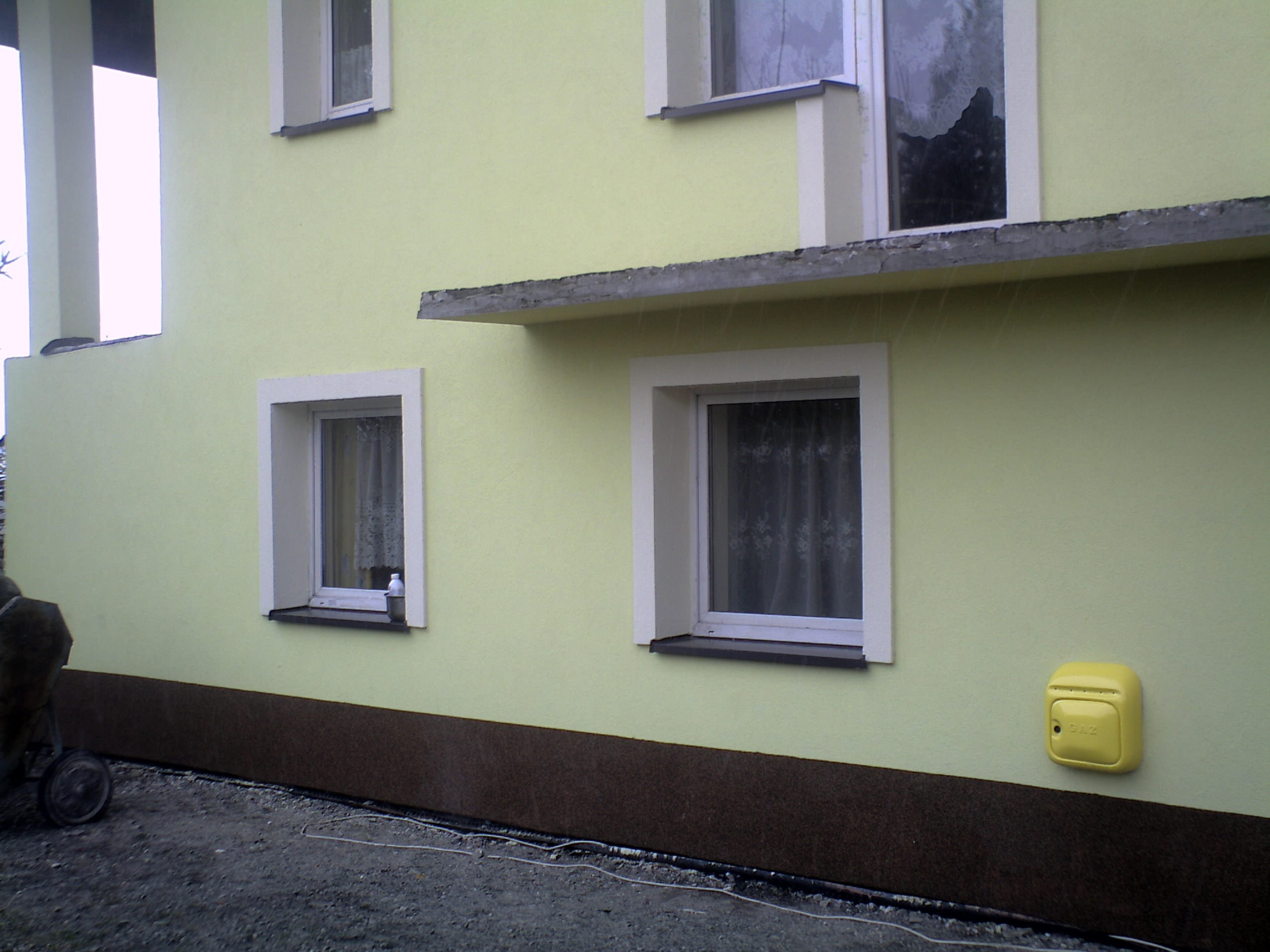 Jasnożółta elewacja domu z białymi obramowaniami okien, brązowym cokołem i żółtą skrzynką gazową, sfotografowana w pochmurny dzień.