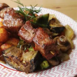 Szczegółowe ujęcie szaszłyka z grillowanego mięsa z warzywami, w tym bakłażanem, papryką i pieczarkami, udekorowanego gałązką rozmarynu, podane na talerzu w geometryczny wzór.