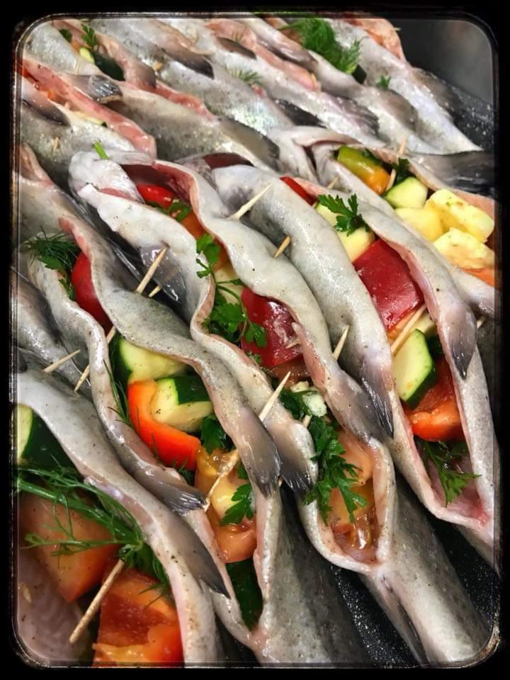 Rząd surowych pstrągów wypełnionych warzywami (papryka, cukinia, ziemniaki, zioła) i spiętych wykałaczkami, gotowych do pieczenia w metalowej brytfannie.