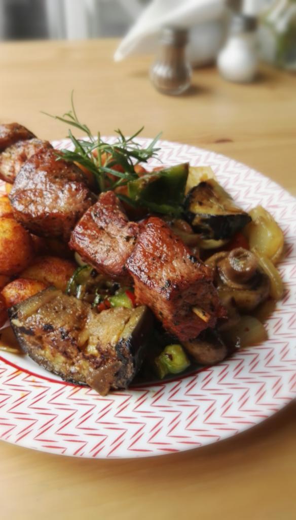 Szczegółowe ujęcie szaszłyka z grillowanego mięsa z warzywami, w tym bakłażanem, papryką i pieczarkami, udekorowanego gałązką rozmarynu, podane na talerzu w geometryczny wzór.