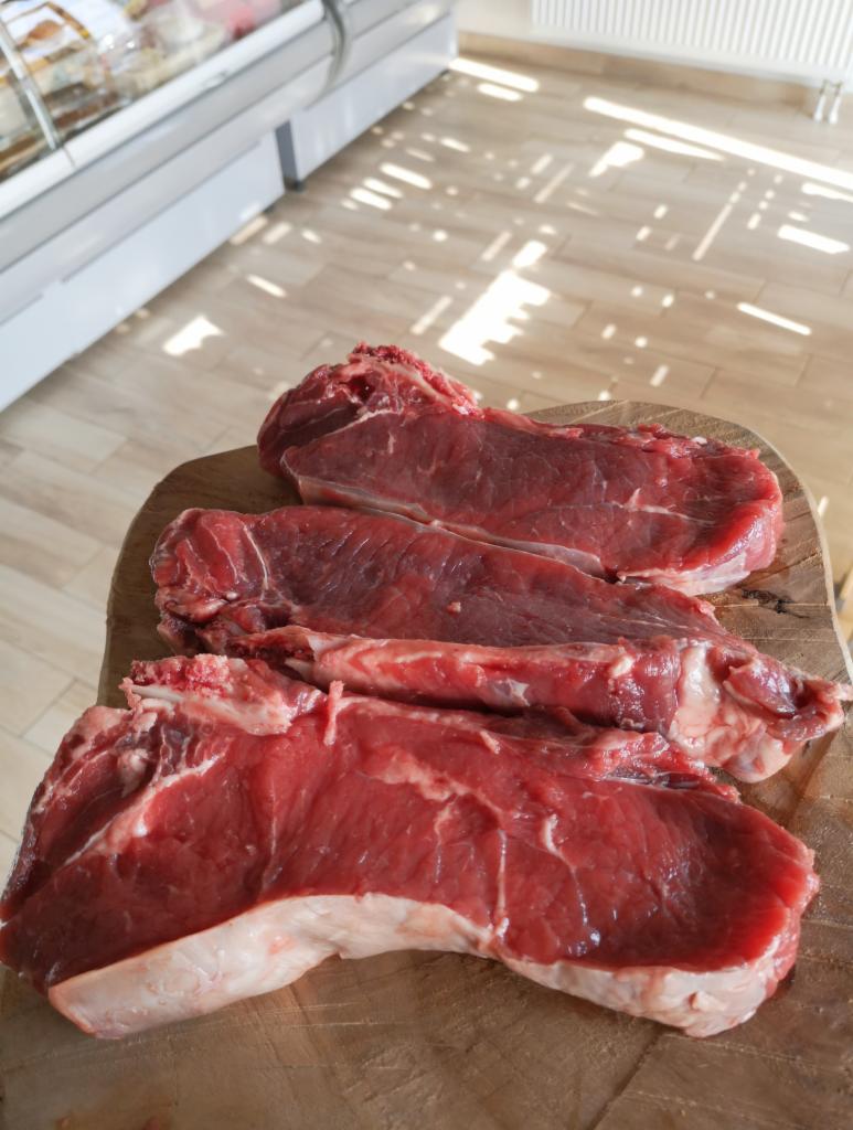 Trzy surowe steki z antrykotu z marmurkowym tłuszczem, ułożone na drewnianej desce, w tle lada chłodnicza i podłoga z jasnych paneli, oświetlone promieniami słonecznymi.