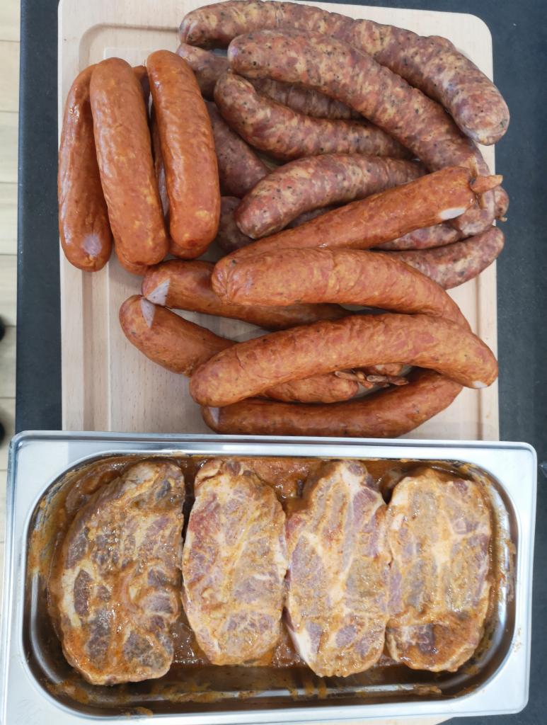 Przygotowane do pieczenia karkówki w marynacie obok różnego rodzaju surowych kiełbas na desce z jasnego drewna.