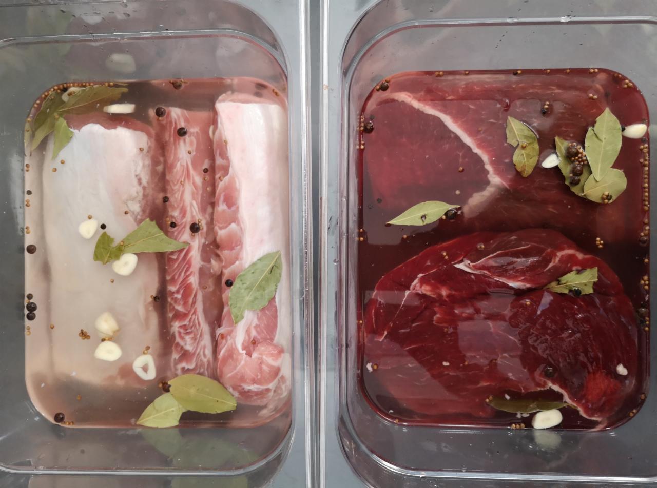 Dwa pojemniki z marynowanym mięsem: wieprzowina i wołowina, z dodatkiem liści laurowych, ziaren pieprzu i czosnku, przygotowywane do pieczenia lub wędzenia.