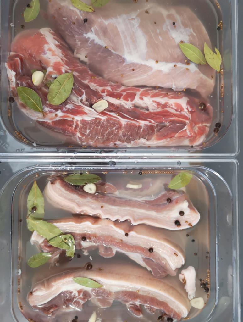 Dwa pojemniki z surowym, peklowanym mięsem wieprzowym w zalewie z liśćmi laurowymi, czosnkiem i ziarnami pieprzu, widoczne z góry.