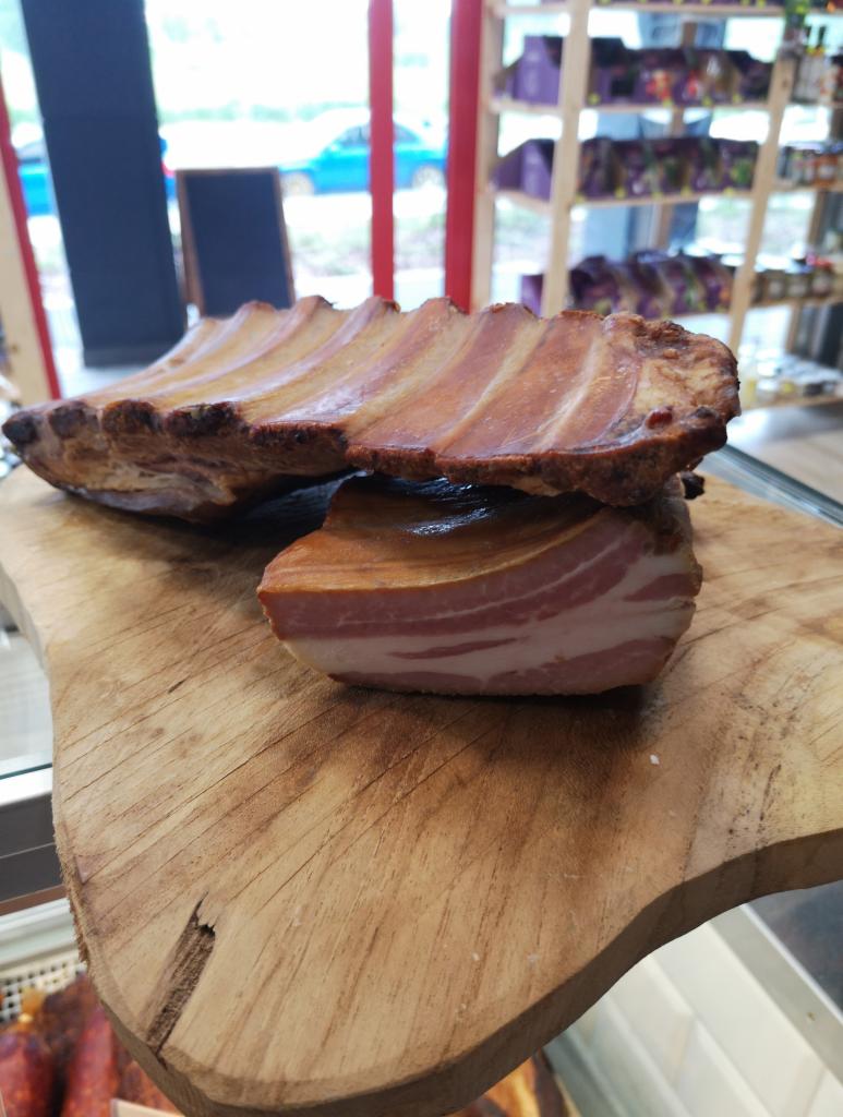 Wędzone żeberka i kawałek boczku na drewnianej desce, widoczne w sklepie za szybą lady chłodniczej.