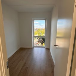 Twins Technologie Wykańczania Wnętrz Szczepan Ścibor - Pusty pokój z drewnianą podłogą i świeżo pomalowanymi białymi ścianami, widok z otwartych drzwi na okno z widokiem na zieleń i budynki.