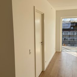 Twins Technologie Wykańczania Wnętrz Szczepan Ścibor - Jasne, świeżo pomalowane wnętrze z drewnianą podłogą i białymi drzwiami, widok na balkon z metalową balustradą i budynkiem w tle.