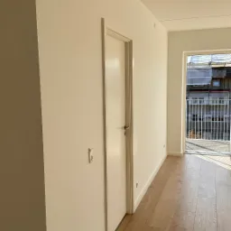 Jasne, świeżo pomalowane wnętrze z drewnianą podłogą i białymi drzwiami, widok na balkon z metalową balustradą i budynkiem w tle.