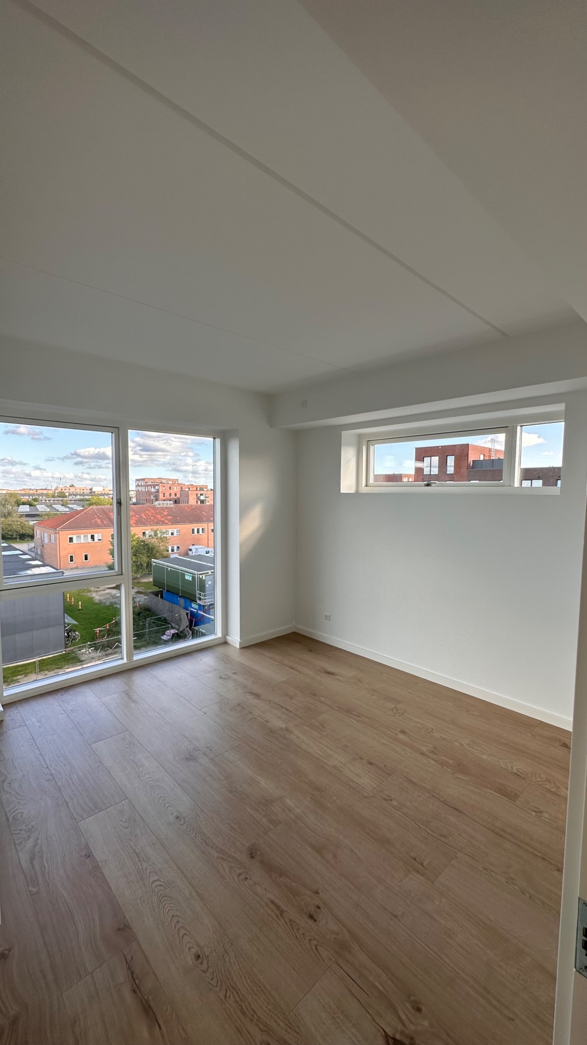 Jasne, puste pomieszczenie z nowymi panelami podłogowymi, świeżo pomalowanymi ścianami i widokiem na budynki z zewnątrz przez okno.