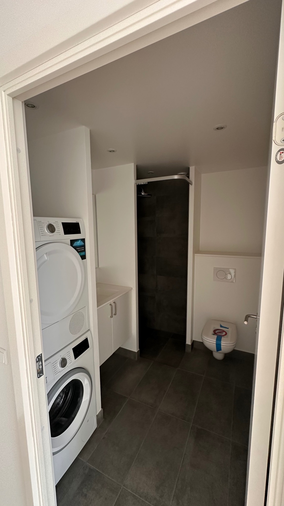 Nowoczesna łazienka z pralką i suszarką w zabudowie, ciemna glazura pod prysznicem, toaleta z niebieską taśmą zabezpieczającą, widok zza futryny drzwi.