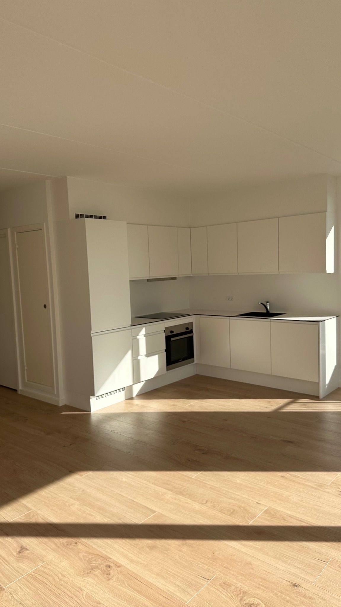 Jasne wnętrze kuchni z białymi meblami w minimalistycznym stylu, drewniana podłoga ułożona w jodełkę, oświetlona promieniami słonecznymi wpadającymi przez okno.