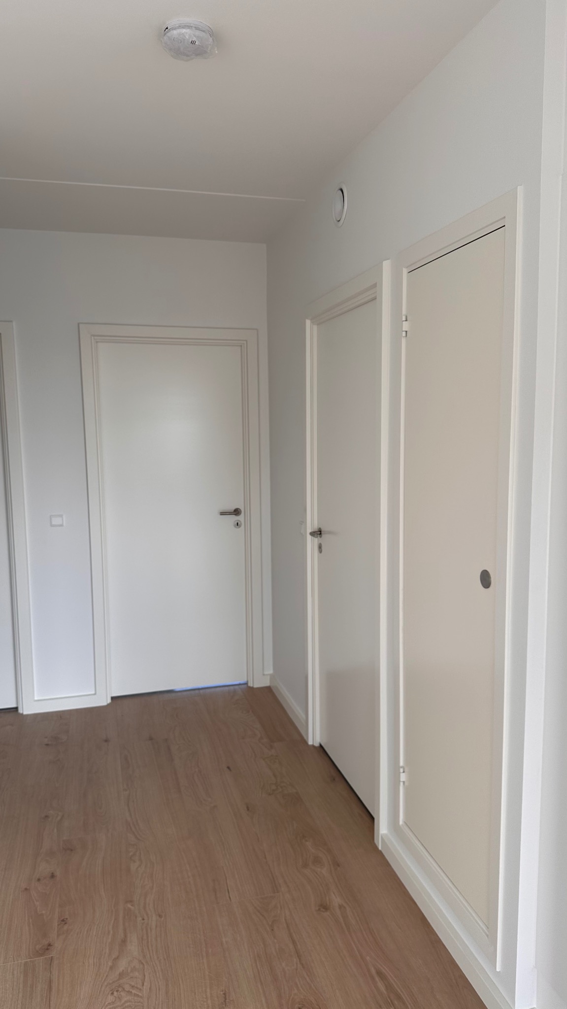 Korytarz z trzema białymi drzwiami i panelami podłogowymi w odcieniach brązu. Na suficie widoczny czujnik dymu w folii.