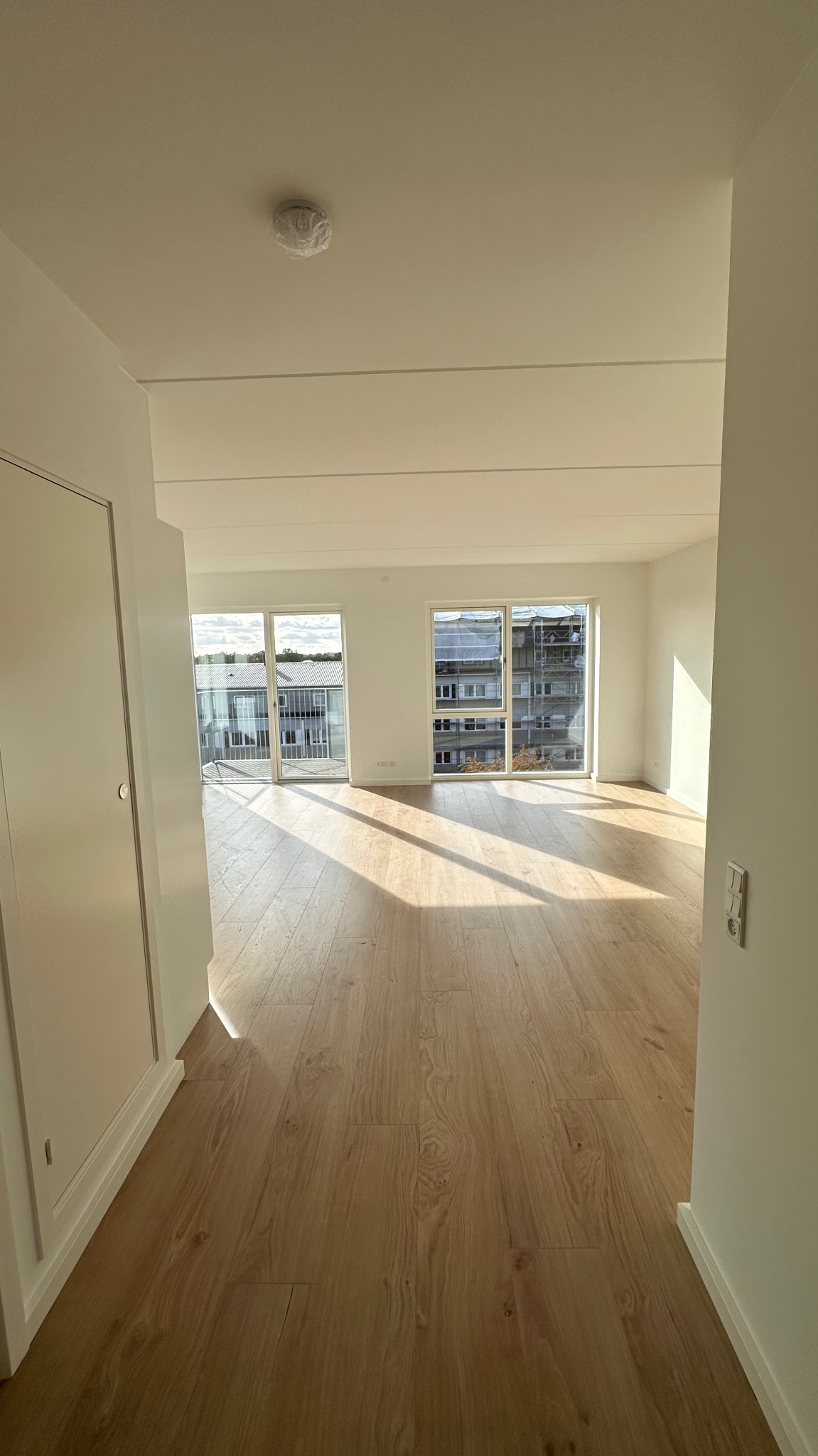 Jasne, przestronne wnętrze mieszkania z drewnianą podłogą i białymi ścianami, widok z korytarza w stronę okien z widokiem na budynek mieszkalny.