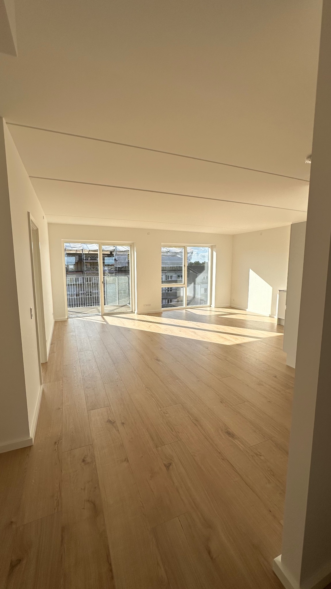 Jasne panele podłogowe w nowym, pustym mieszkaniu z białymi ścianami i dużymi oknami balkonowymi, wpadające promienie słoneczne.