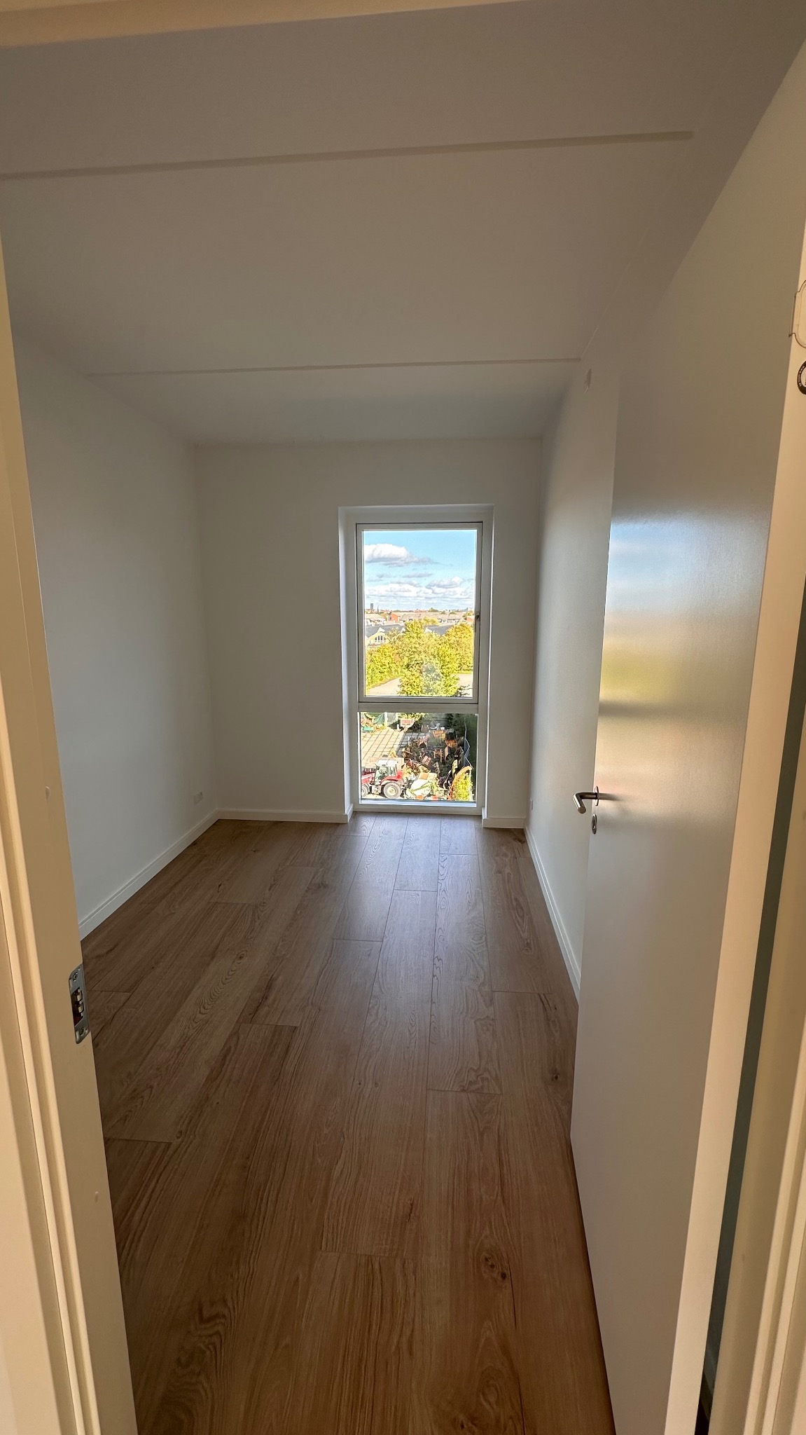 Pusty pokój z drewnianą podłogą i świeżo pomalowanymi białymi ścianami, widok z otwartych drzwi na okno z widokiem na zieleń i budynki.