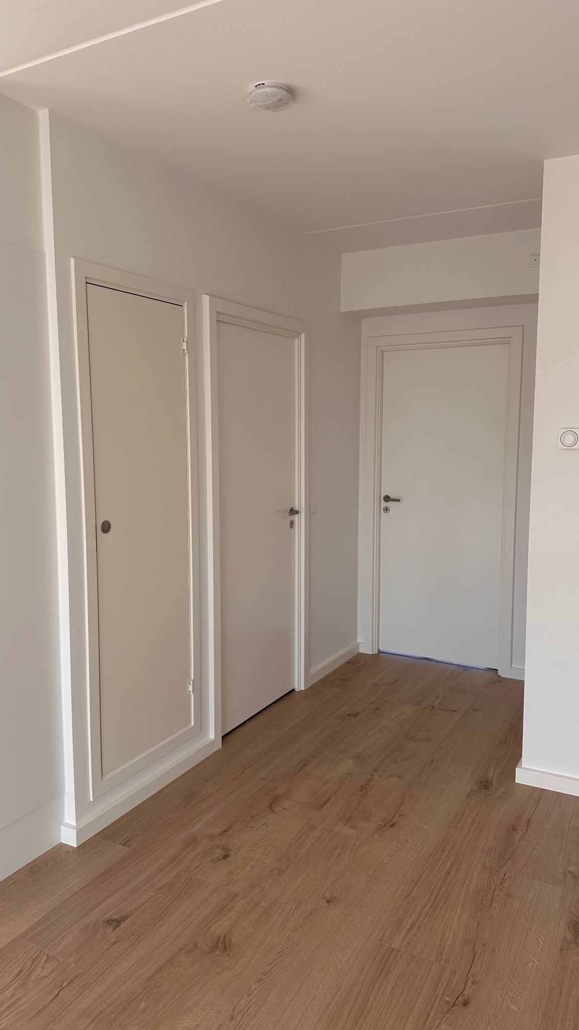 Jasne, nowocześnie wykończone wnętrze z trzema białymi drzwiami, podłogą z paneli imitujących drewno i czujnikiem dymu na suficie.