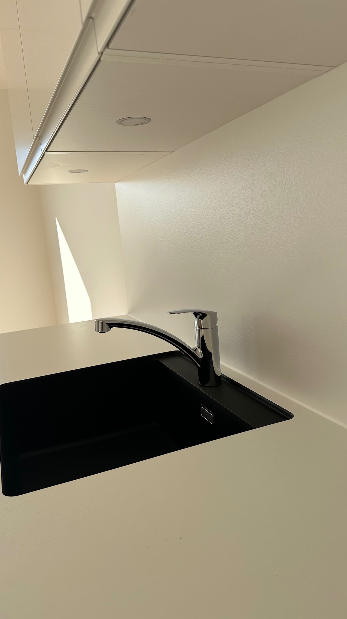 Minimalistyczna kuchnia: czarny zlew z chromowaną baterią na tle jasnej ściany i szafek z oświetleniem punktowym.