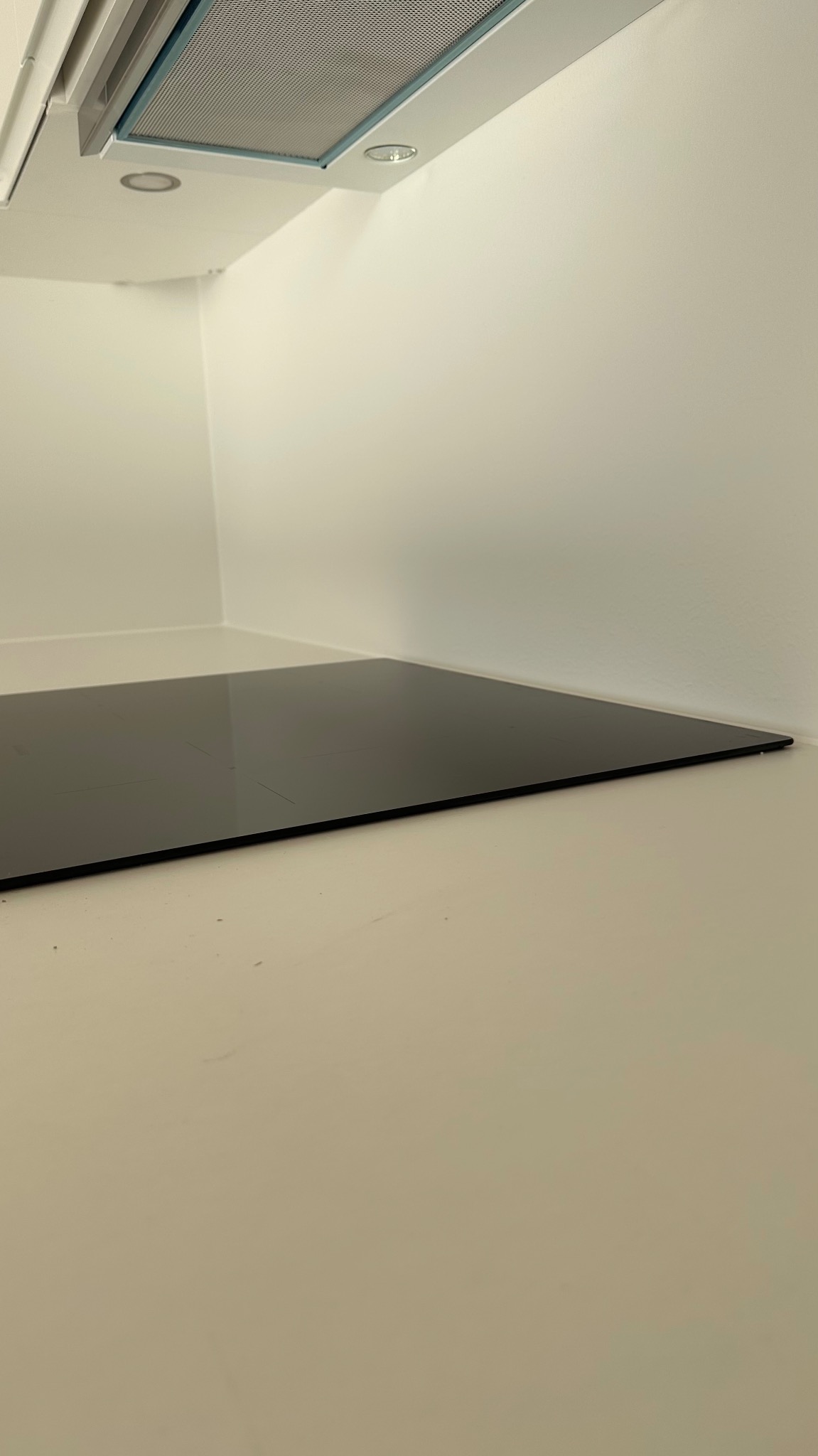 Minimalistyczna kuchnia z czarną płytą indukcyjną wpuszczoną w jasny blat, widok z niskiego kąta, oświetlenie sufitowe i białe ściany.