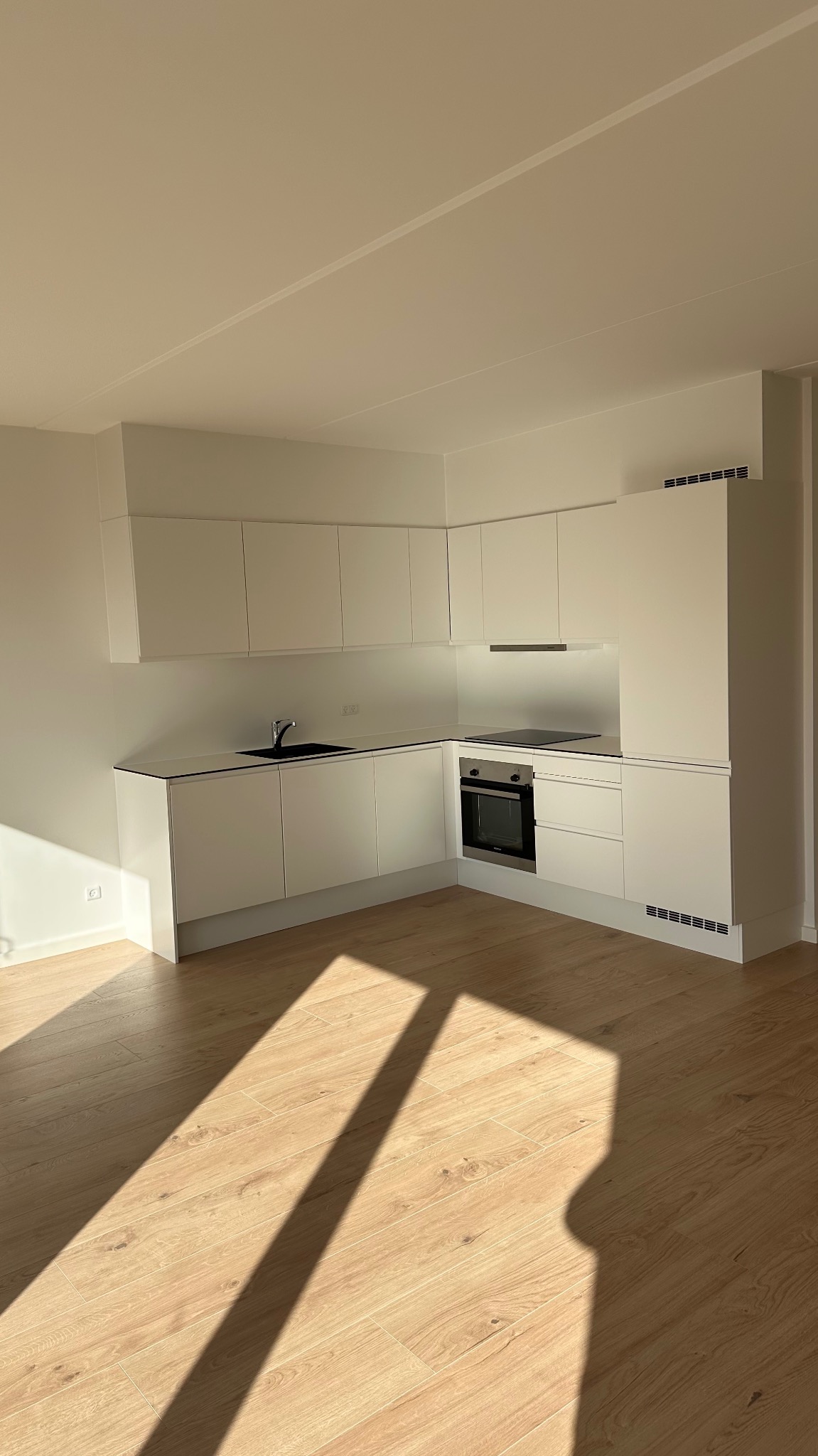 Jasna, nowoczesna kuchnia z białymi szafkami i drewnianą podłogą, zalana popołudniowym słońcem tworzącym wyraźne cienie.