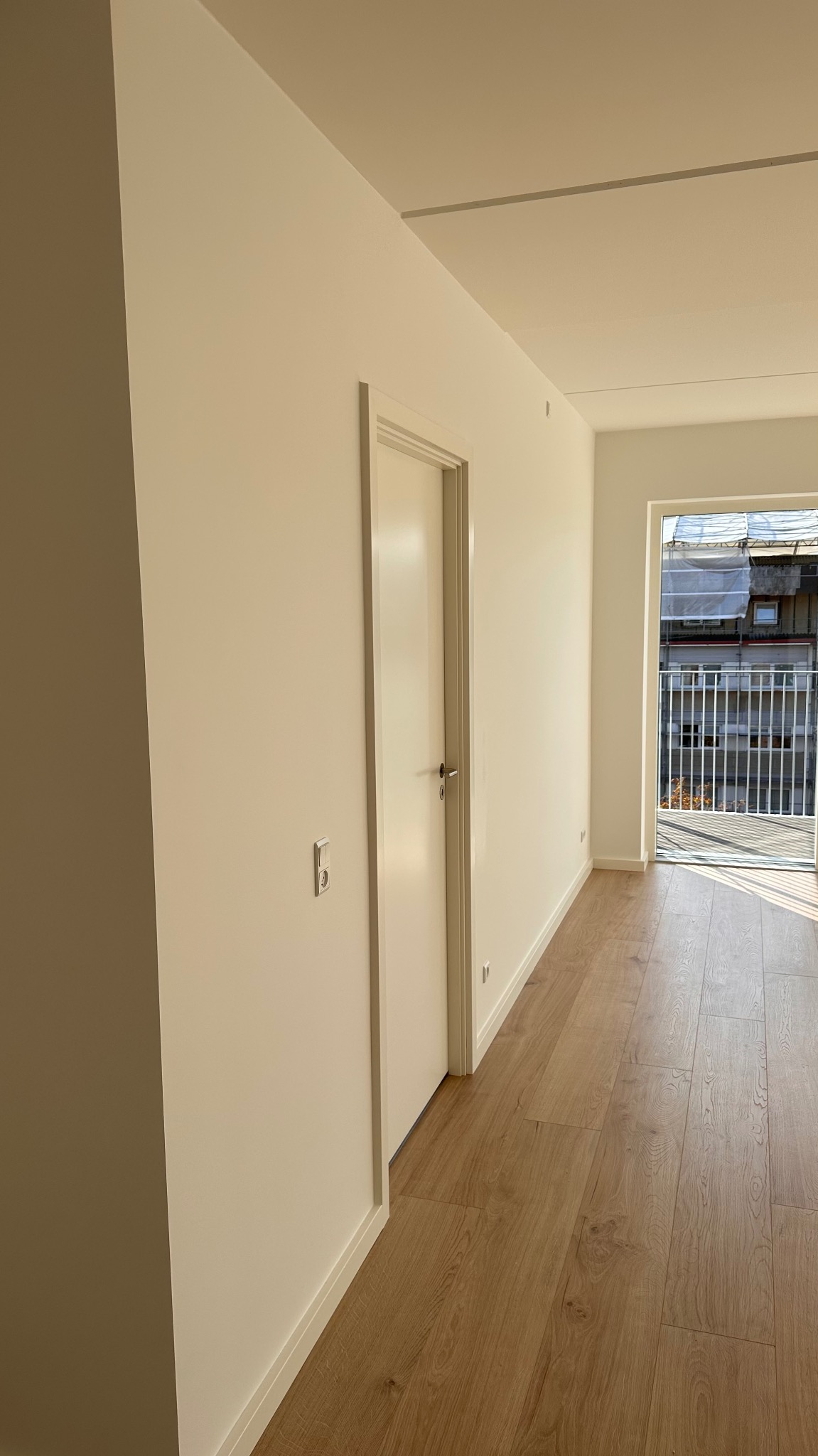 Jasne, świeżo pomalowane wnętrze z drewnianą podłogą i białymi drzwiami, widok na balkon z metalową balustradą i budynkiem w tle.