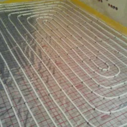 Białe rury ogrzewania podłogowego ułożone w pętle na srebrnej macie z czerwoną siatką, widok z góry na niedokończoną instalację.