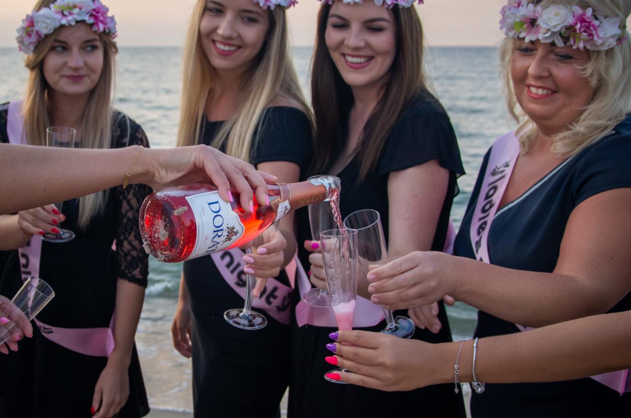 Grupa kobiet w wiankach i szarfach 'Ladies Night' świętuje na plaży, nalewając różowe wino musujące do kieliszków.