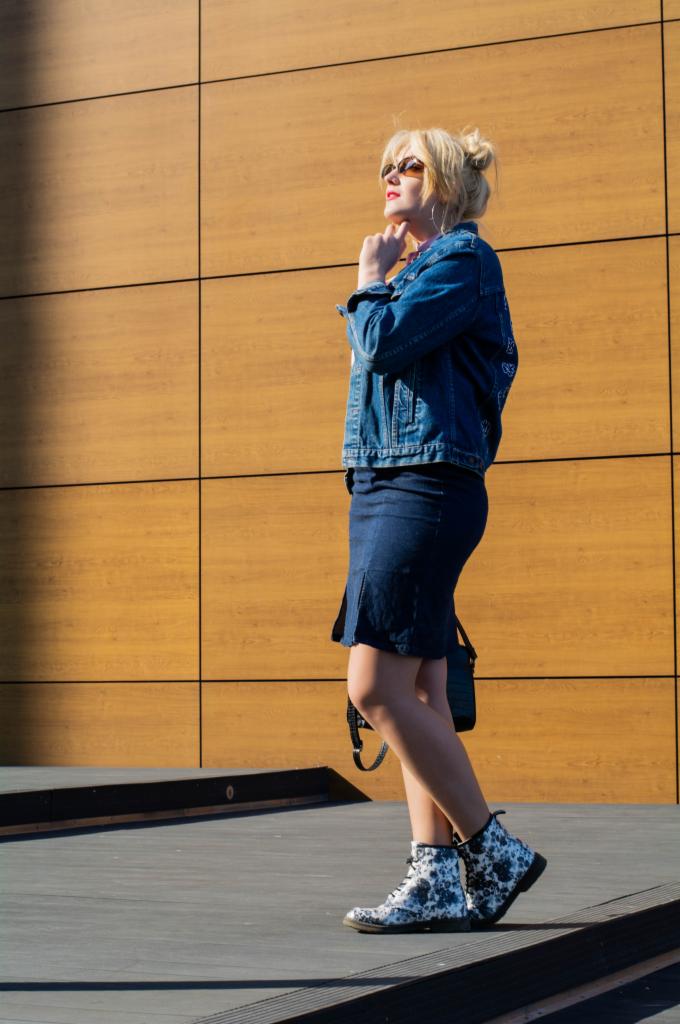 Kobieta w dżinsowej kurtce i spódnicy, z blond włosami upiętymi w kok, pozuje na tle drewnianej ściany, ubrana w wzorzyste botki i okulary przeciwsłoneczne.