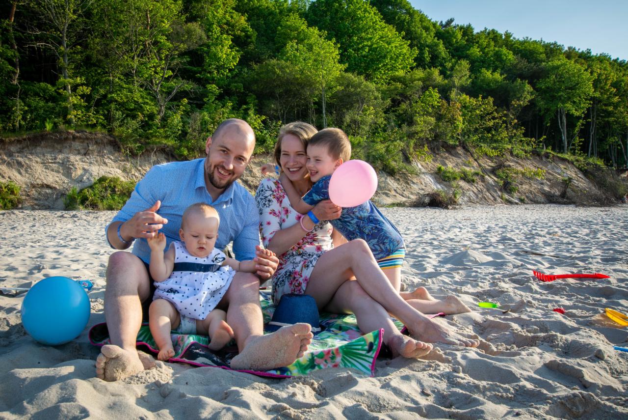 Uśmiechnięta rodzina z dwójką dzieci pozuje na piaszczystej plaży z klifowym brzegiem w tle. Dziecko trzyma różowy balon.