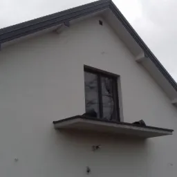 Boczny widok na nowo otynkowany dom z ciemnymi rynnami i oknem na poddaszu, widoczne elementy instalacji na elewacji i iglaste drzewo w tle.