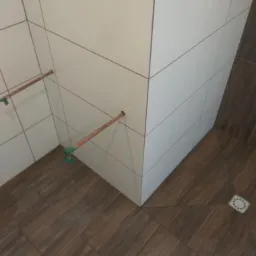 Świeżo ułożone, białe, prostokątne płytki na ścianie w rogu łazienki, z wystającymi rurami miedzianymi zabezpieczonymi zieloną taśmą, oraz ciemnobrązowe płytki imitujące drewno na podłodze...