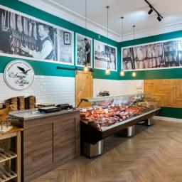 IG Design - Wnętrze sklepu mięsnego z ladą chłodniczą pełną wędlin, drewnianą ścianą z geometrycznym wzorem oraz czarno-białymi fotografiami na ścianach w kolorze butelkowej zieleni.