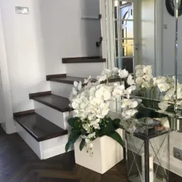 Schody z ciemnego drewna z białymi podstopnicami, dekorowane białymi orchideami w kwadratowej donicy i lustrzaną latarnią, parkiet w jodełkę.