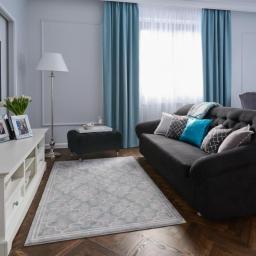 IG Design - Salon w odcieniach szarości i błękitu: ciemnoszara sofa z dekoracyjnymi poduszkami, wzorzysty dywan na parkiecie w jodełkę, biała komoda z ramkami na zdjęcia i tulipanami, turkusowe zasłony.