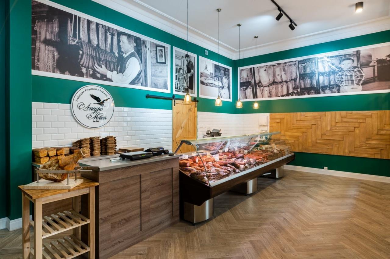 Wnętrze sklepu mięsnego z ladą chłodniczą pełną wędlin, drewnianą ścianą z geometrycznym wzorem oraz czarno-białymi fotografiami na ścianach w kolorze butelkowej zieleni.