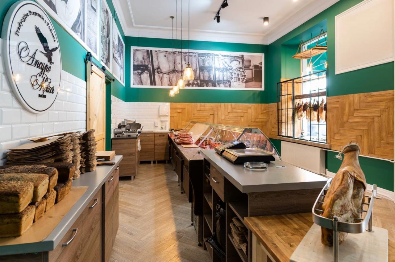 Wnętrze sklepu z zielonymi ścianami, drewnianymi elementami i ekspozycją pieczywa oraz wędlin w chłodniach.