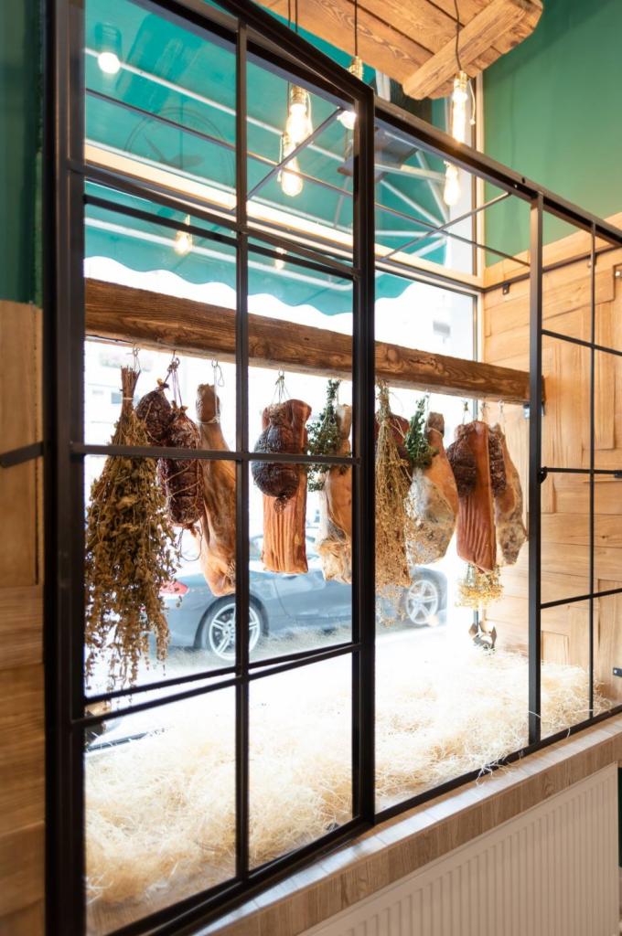 Nowoczesna witryna z czarnymi szprosami, za którą wisi suszone mięso i zioła na tle ulicy z zaparkowanym srebrnym samochodem.