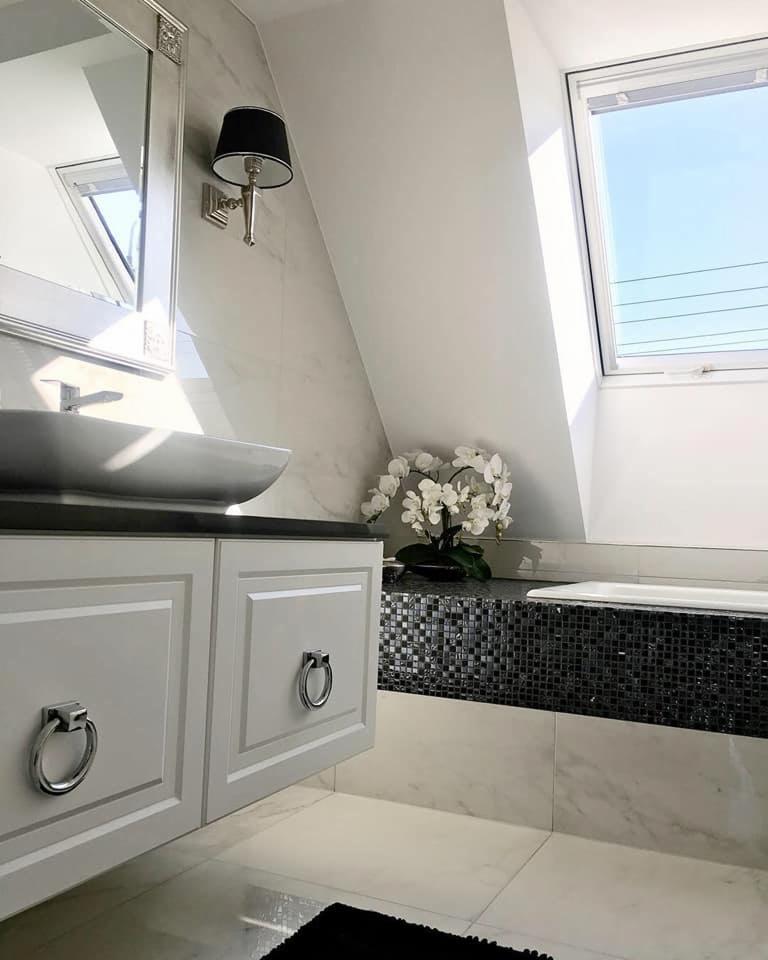 Elegancka łazienka na poddaszu z białą armaturą, umywalką nablatową, szafką z frezowanymi frontami i okrągłymi uchwytami, mozaiką przy wannie i storczykiem.