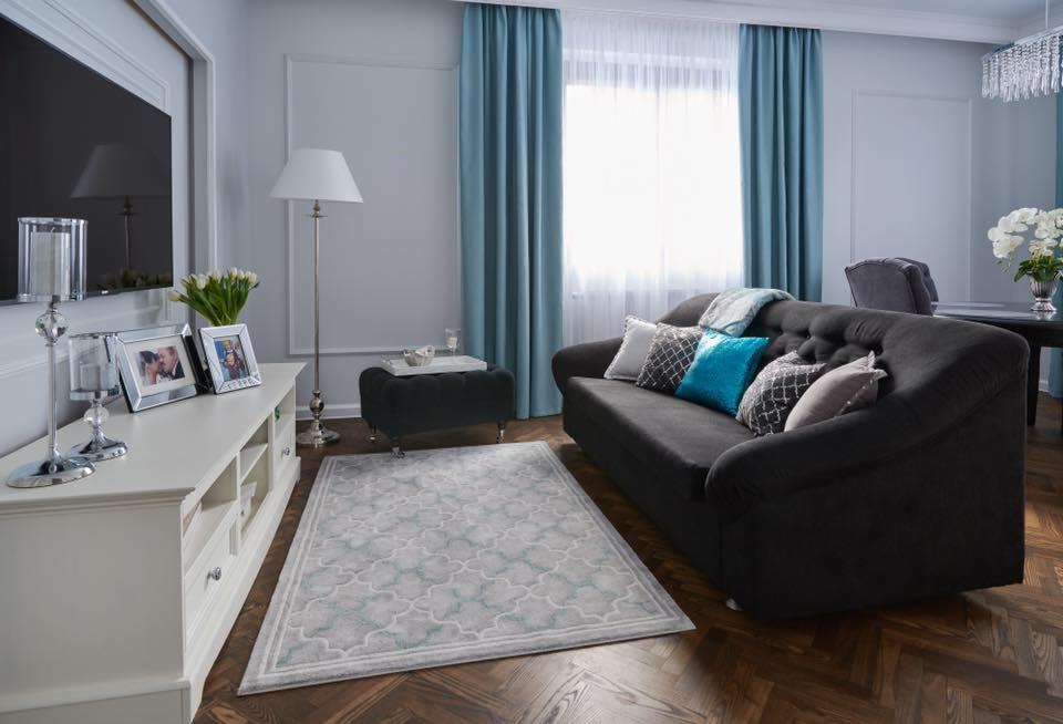 Salon w odcieniach szarości i błękitu: ciemnoszara sofa z dekoracyjnymi poduszkami, wzorzysty dywan na parkiecie w jodełkę, biała komoda z ramkami na zdjęcia i tulipanami, turkusowe zasłony.