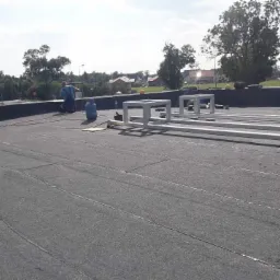 Prace na płaskim dachu pokrytym papą, widoczne metalowe konstrukcje wsporcze oraz butle gazowe, dekarz w oddali, w tle zabudowa jednorodzinna i drzewa.