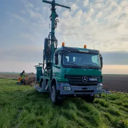Wiertnica studni głębinowej zamontowana na zielonym samochodzie ciężarowym Mercedes Actros, obok pracuje koparka wykopująca ziemię, pole w tle.