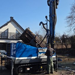 WATEKO TOMASZ PYCHYŃSKI - Wiertnica ROTOMAX XL GTC w trakcie pracy przy budowie studni głębinowej. Operator w kombinezonie obsługuje maszynę na tle domu i błękitnego nieba.