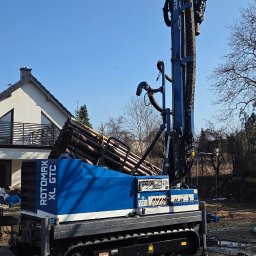 WATEKO TOMASZ PYCHYŃSKI - Niebieska wiertnica Rotomax XL GTC na gąsienicach, z rurami wiertniczymi, na tle domu i błękitnego nieba. Sprzęt do wiercenia studni.