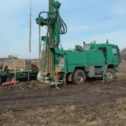 Zielona ciężarówka z wiertnicą na polu, widoczne rury wiertnicze na przyczepie, ślady błota na ziemi.