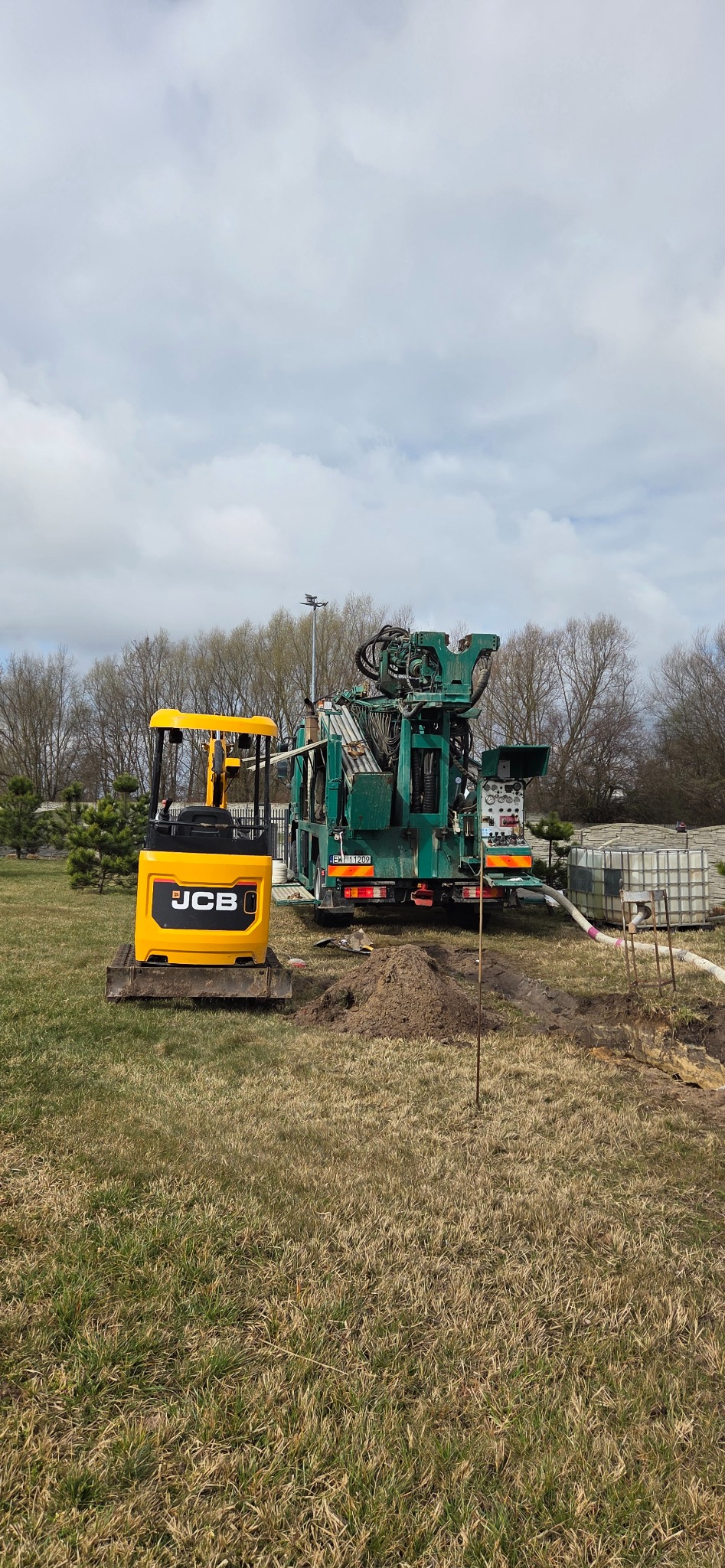 Żółta mini koparka JCB i wiertnica na trawiastym terenie. W tle drzewa i zbiorniki na wodę. Widok na sprzęt podczas wiercenia studni.