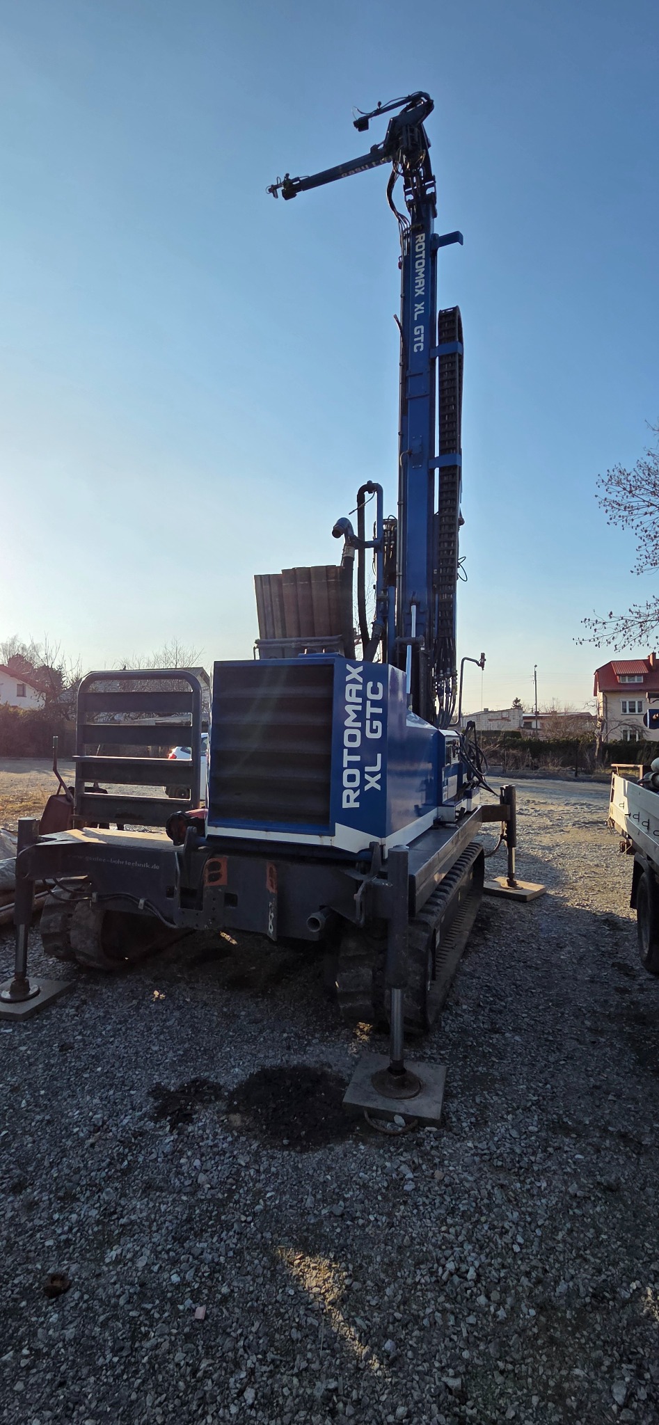 Niebieska wiertnica Rotomax XL GTC na żwirowym podłożu, z wysuniętym ramieniem w górę, na tle błękitnego nieba i zabudowań.