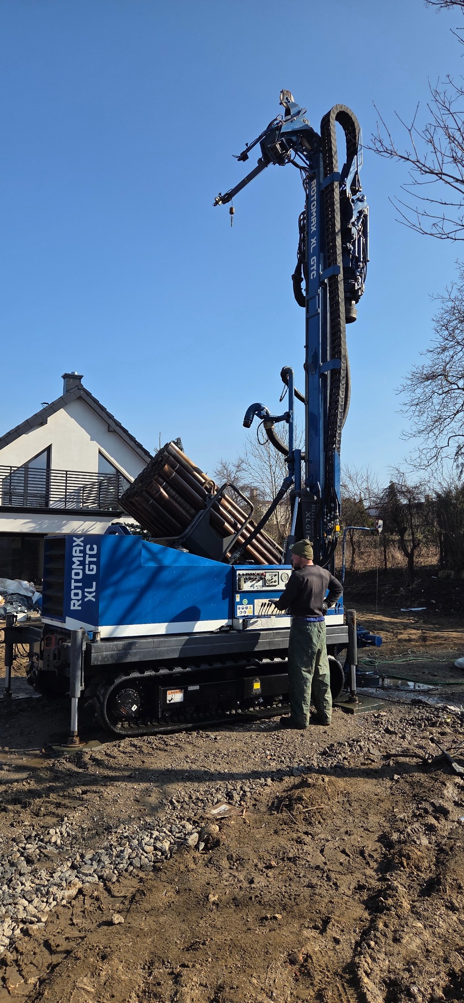 Wiertnica ROTOMAX XL GTC w trakcie pracy przy budowie studni głębinowej. Operator w kombinezonie obsługuje maszynę na tle domu i błękitnego nieba.