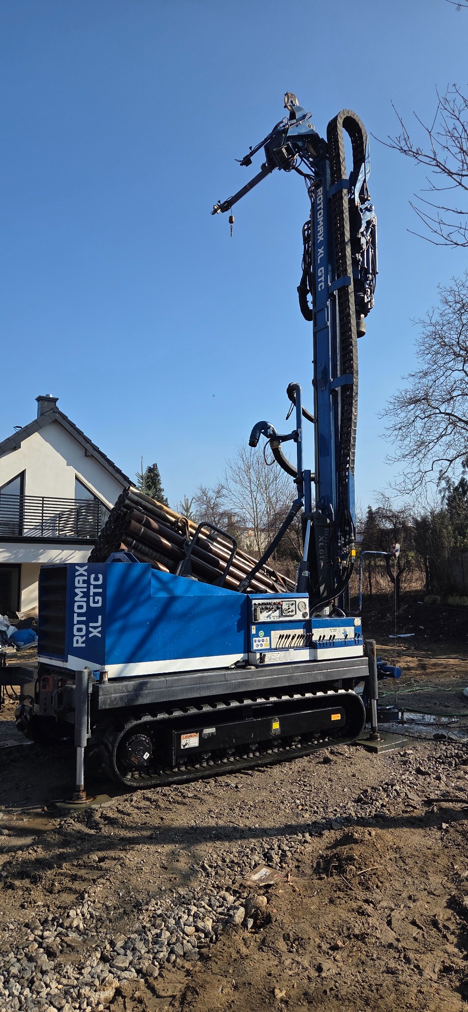Niebieska wiertnica Rotomax XL GTC na gąsienicach, z rurami wiertniczymi, na tle domu i błękitnego nieba. Sprzęt do wiercenia studni.