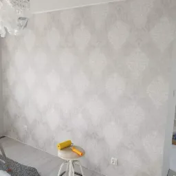 Ściana po tapetowaniu z delikatnym, symetrycznym wzorem w odcieniach szarości, widoczna drabina malarska i wałek do tapet na stołku.