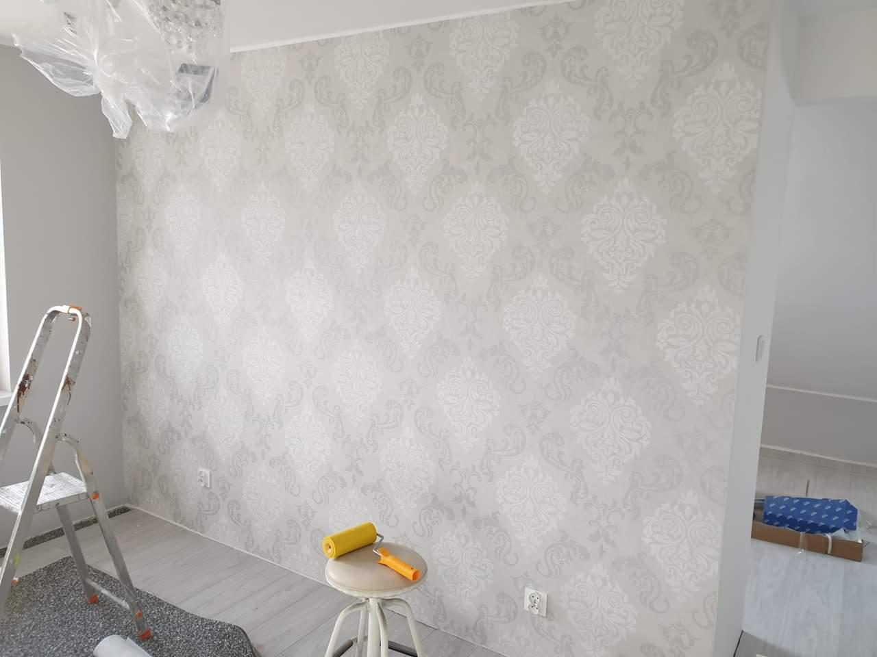 Ściana pokryta tapetą w delikatne, jasnoszare wzory, z drabinką malarską i wałkiem do tapet na stołku w pomieszczeniu w trakcie remontu.